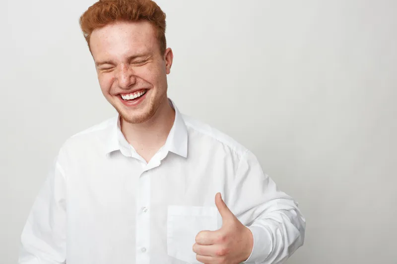 Happy young man with red hair and white shirt smiles with eyes closed showing thumbs up gesture on light background. Concept of positivity, approval, and confidence.
