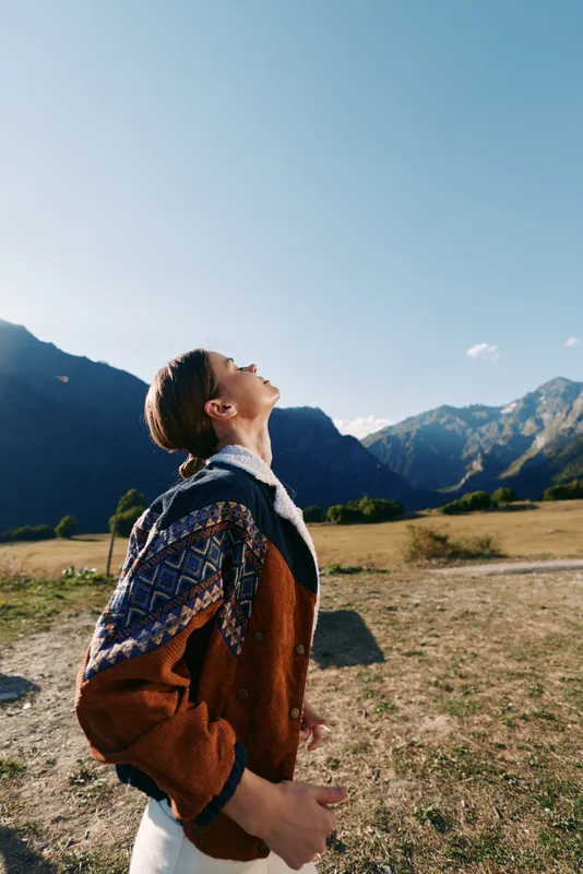 Woman hiking with backpack in mountains and nature, outdoors travel scene. Sweater-clad female looking up at blue sky on sunny day, landscape adventure and exploration in wilderness.