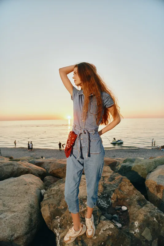 lifestyle woman in modern boho-western denim street style posing on rocks by ocean at sunset with film color tones and relaxed casual vibe