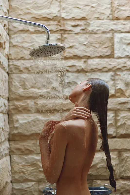 woman taking shower under rainfall showerhead with wet hair and stone wall background, relaxing self-care moment in warm light, natural skin and calm mood