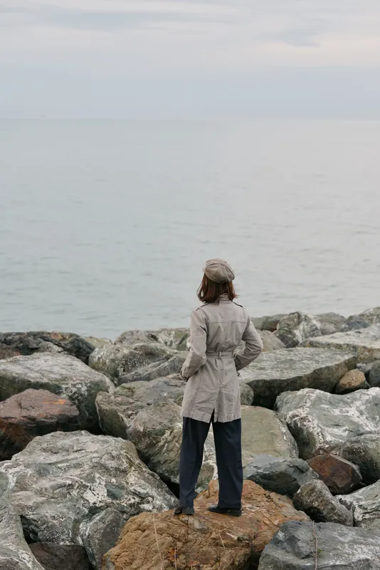 A woman wearing a coat and hat stands on rocky shore facing the calm sea, contemplative mood on an overcast day. Concept of solitude, nature, and quiet reflection.