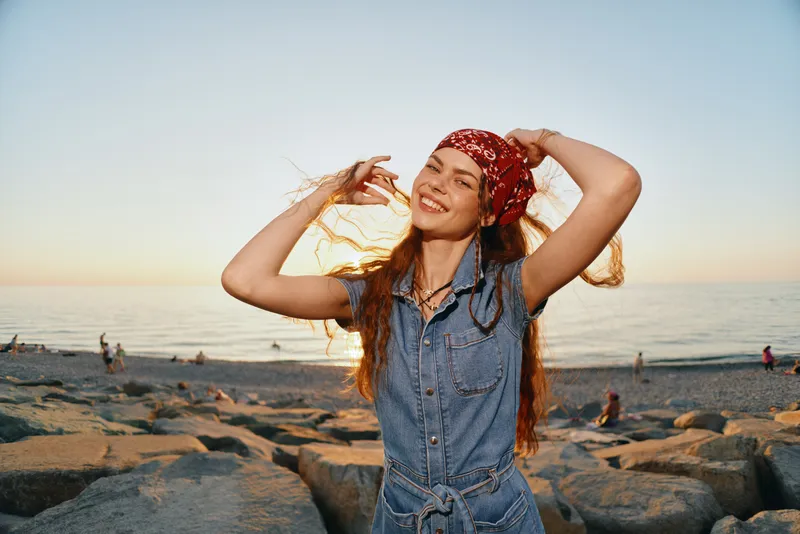 smiling woman in denim jumpsuit and red bandana enjoys sunset at rocky beach with calm sea and warm light, expressing happiness and freedom outdoors