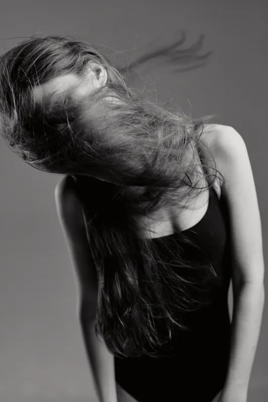Black and white portrait of a woman with long hair flowing, wearing a black sleeveless top, expressing motion and emotion.
