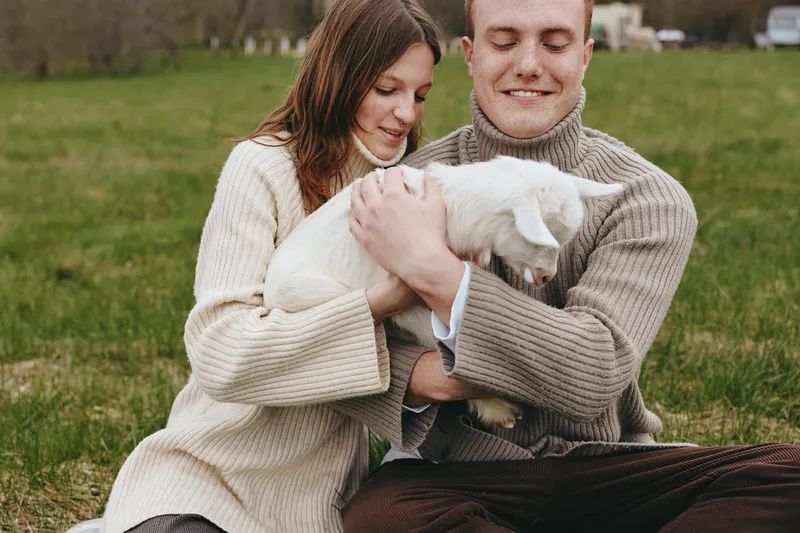 Smiling couple cuddles a white goat kid in a grassy field, cozy sweaters, warm light. Lifestyle scene highlighting connection with animals, nature, and simple living. Tender and peaceful moment