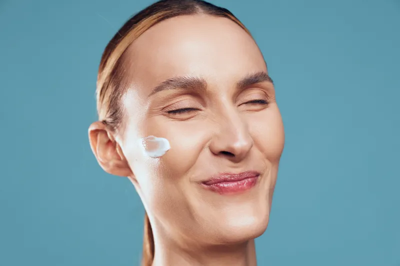Closeup of woman with cream on cheek smiling with eyes closed, skincare routine, fresh natural look, smooth skin texture, blue background, wellness concept