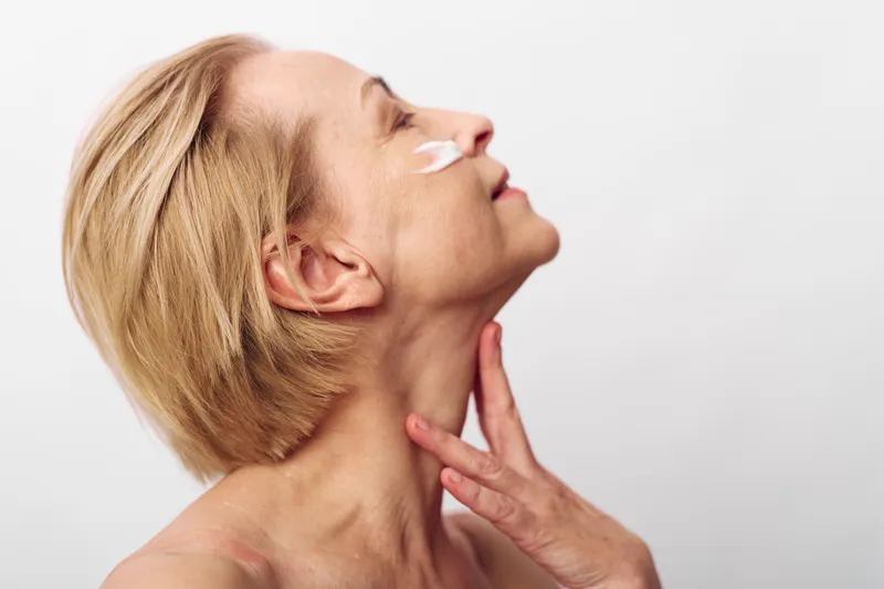 Blonde middle aged woman applies white cream on cheek with closed eyes and relaxed expression. Skin care concept for beauty, wellness, anti aging, and facial hydration in studio light.