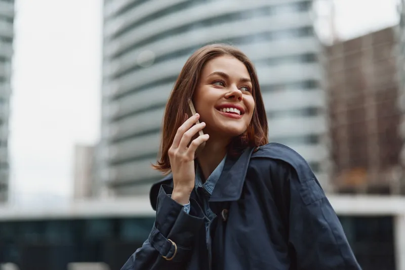 Young woman in dark coat talks on smartphone outdoors in urban environment smiles happily enjoys conversation during daytime city life background modern buildings.