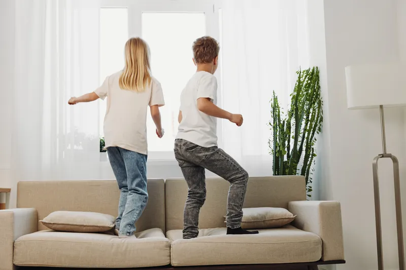 Two children jumping on a beige sofa in a bright living room with natural light and green plant. Concept of playful energy, childhood fun, and home activity.
