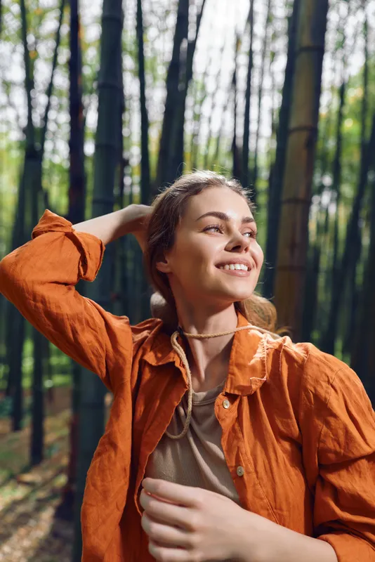 Woman smile bamboo forest outdoor nature portrait of a young female in orange jacket, sunlight on her face as she looks upward, relaxed and joyful expression in a serene green grove.