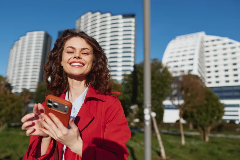 A smiling young woman in a red jacket holds a smartphone outdoors with modern high rises behind her, capturing a candid moment of everyday urban life on a bright day.