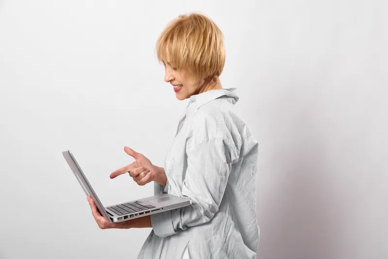 Woman with short blonde hair uses laptop for typing in a bright white office, technology and remote communication concept, focused side view, light clothing, modern work, routine