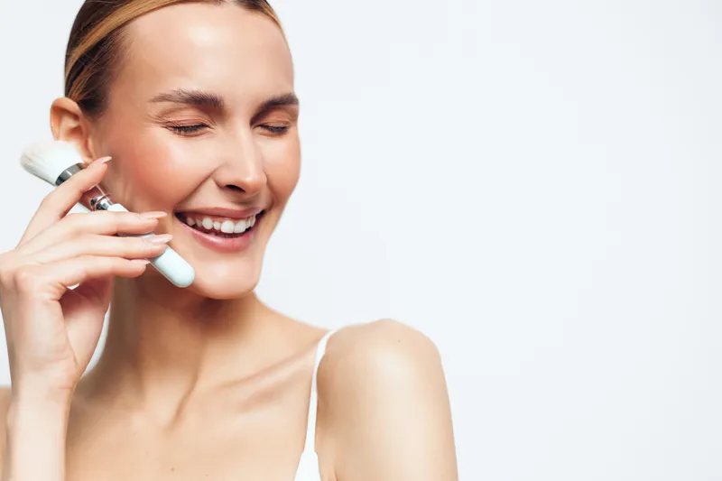 Smiling woman with closed eyes using facial cleansing brush on smooth skin, skincare routine, fresh clean face, white background, beauty concept, self care