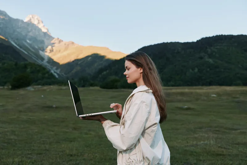 A focused professional woman works on a laptop in an open field with mountains in the distance. She wears a light jacket, creating a calm, productive outdoor work mood.
