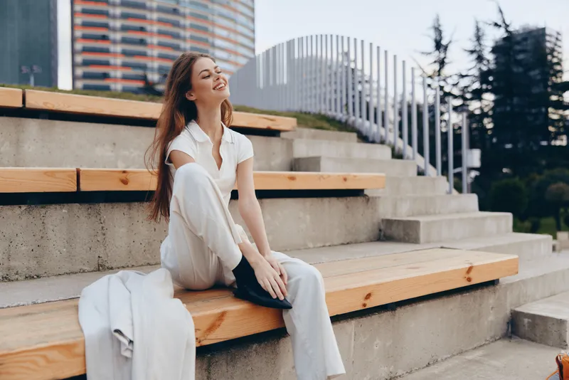 Young woman in white outfit sitting outdoors on wooden steps with joyful smile, urban background, casual fashion and spring mood