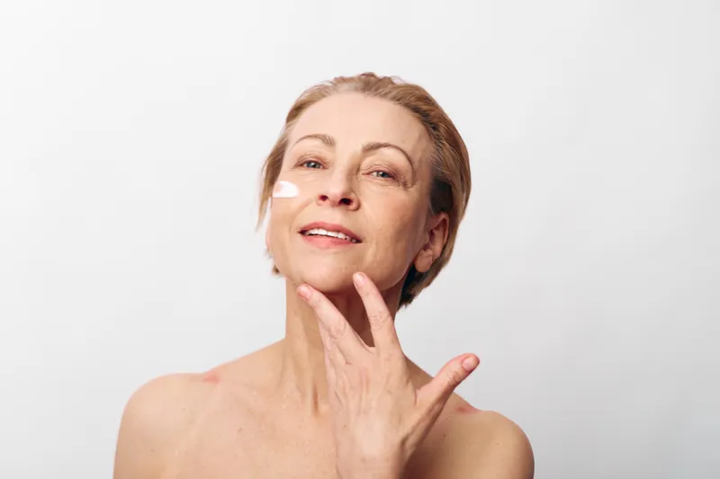 Mature woman with short hair applying cream on face, smiling gently against white background. Concept of skincare, aging beauty, self-care, and natural wellness for mature skin.
