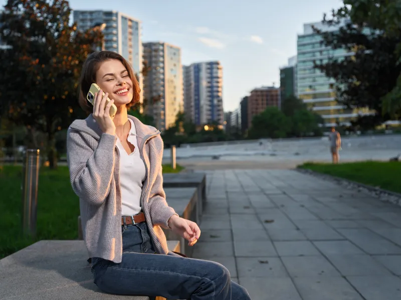 A cheerful professional woman sits outdoors in a modern urban park, smiling while talking on a mobile phone, projecting confidence and a positive, contemporary work vibe.