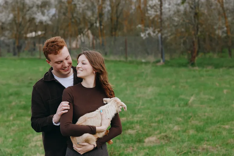 A couple outdoors with a friendly goat kid in a park, sharing a warm moment, smiling at each other, holding the kid, relaxed lifestyle, natural light, candid scene, pastoral atmosphere.