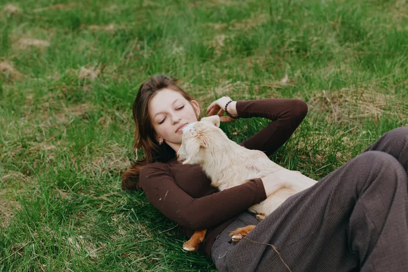Lifestyle moment of a woman relaxing outdoors on grass, gently cuddling a curious goat kid, sunlit scene, soft breeze, natural colors, peaceful expression, casual clothing, wholesome countryside vibe