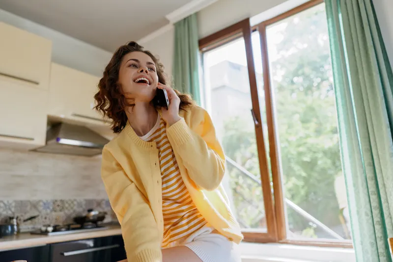 A young woman in a yellow cardigan chats on her phone in a bright kitchen, smiling and relaxed, capturing a casual home moment with sunlight streaming through large windows and mint curtains.