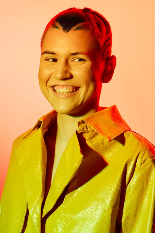 smiling woman with braided hair wearing yellow shiny jacket on warm colorful background, vibrant lighting, joyful expression, portrait close-up