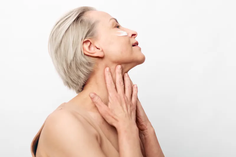 Mature woman with short grey hair applies white cream under eye, gently touching neck on white background. Concept of skincare, aging beauty, self-care, and facial wellness.
