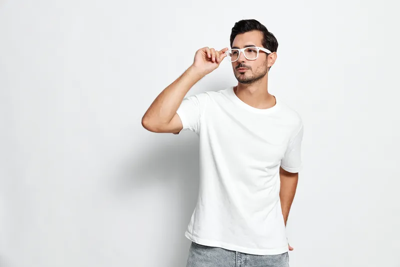 Stylish young man wearing white t-shirt and glasses, looking thoughtful while posing against a plain light background. Minimalist portrait concept.