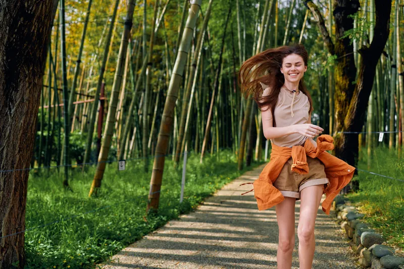 Woman smiling walking along a bamboo forest path enjoying outdoor motion and summer leisure, dressed casual with jacket tied, hair blowing and carefree expression on scenic trail.