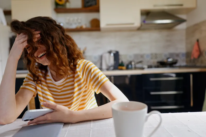 A young woman in a yellow striped shirt sits at a kitchen table, head in her hand, laughing at her phone while a white mug sits in the foreground, capturing a candid moment at home.