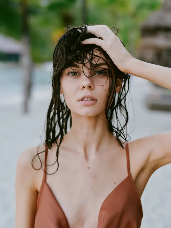 Wet woman with dark hair touching her head outdoors, wearing a brown top, with a natural beach background and relaxed expression.