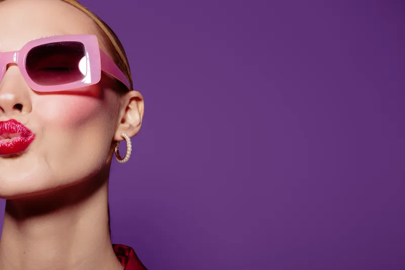 Stylish woman with pink sunglasses and red lipstick blows a kiss on purple background. Close-up portrait with hoop earrings and smooth skin. Concept of fashion, beauty, and glamour.