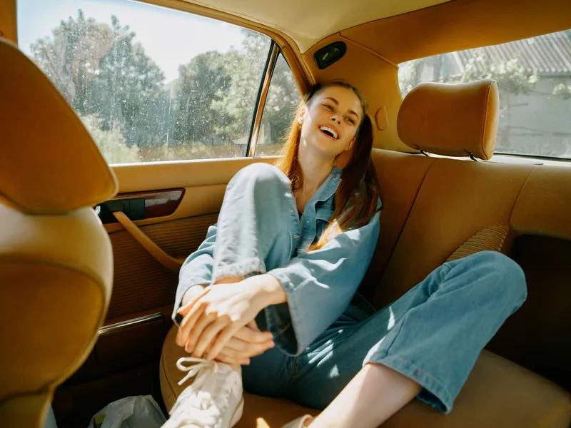 Joyful young woman in casual denim outfit relaxing in the back seat of a car, capturing a carefree moment during a sunny day, representing freedom and adventure.