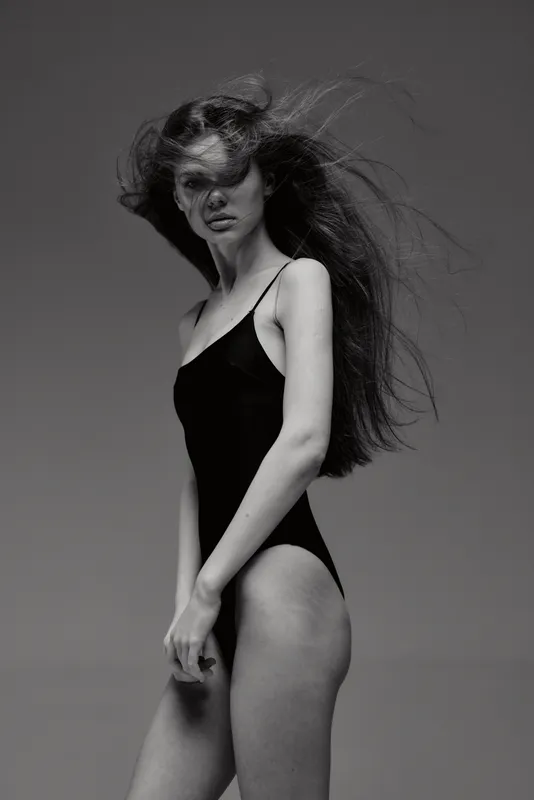 Black and white portrait of a woman with long hair wearing a black dress, posing against a plain background with a dramatic windy effect.