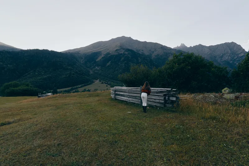 Mountains meadow fence person backpack hiking landscape solitude grass nature. Lone hiker walks beside a wooden fence across a grassy field toward forested mountain peaks under moody sky.