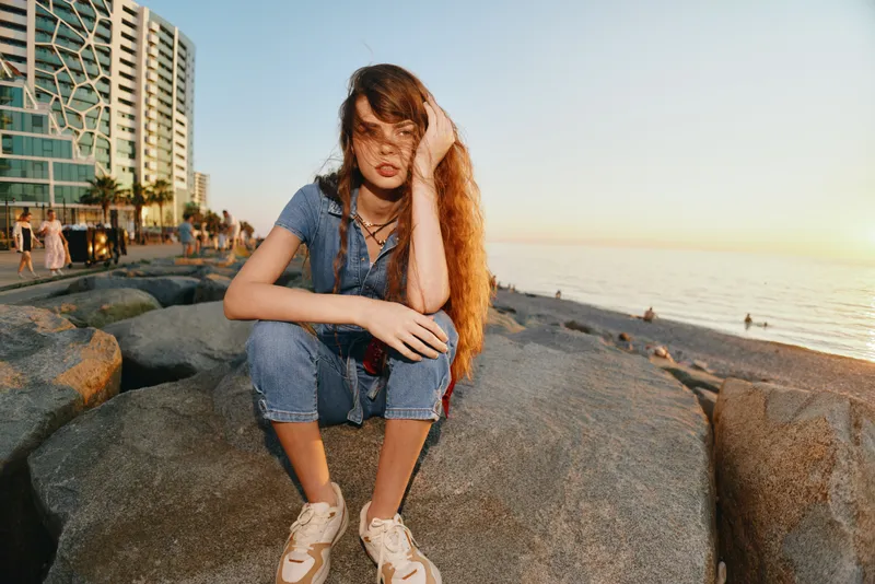 Lifestyle woman in modern boho-western denim street style sitting on rocks by the ocean during golden hour, film color capturing relaxed sunset vibe.