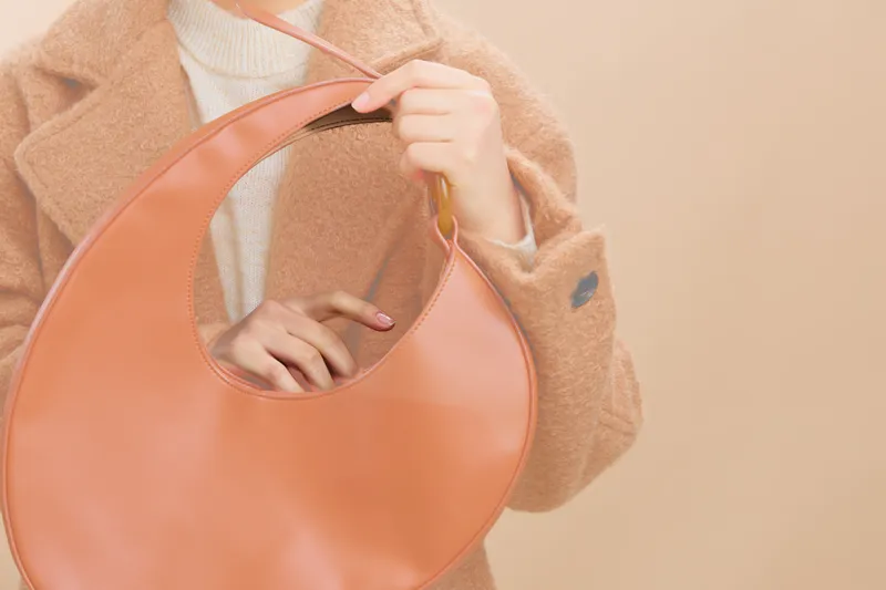asian woman wearing a soft coat holds a round handbag, capturing a chic fashion moment in a muted studio setting. She exudes calm confidence and contemporary style.