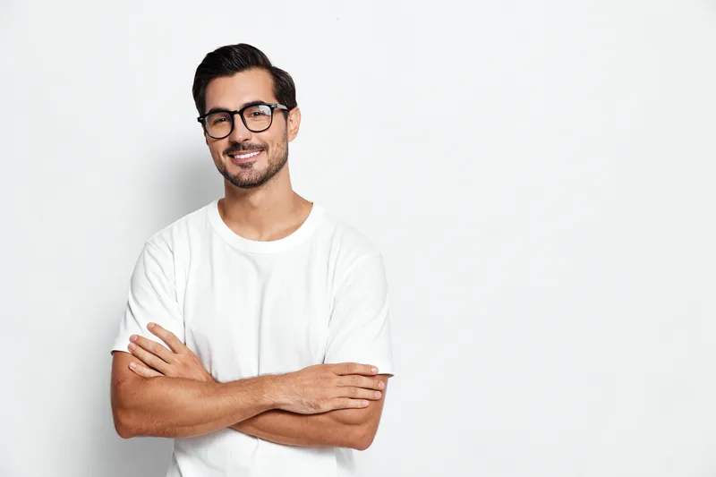Smiling man with glasses standing confidently, wearing a casual white t-shirt, arms crossed in front of a light background, creating a friendly and inviting atmosphere.