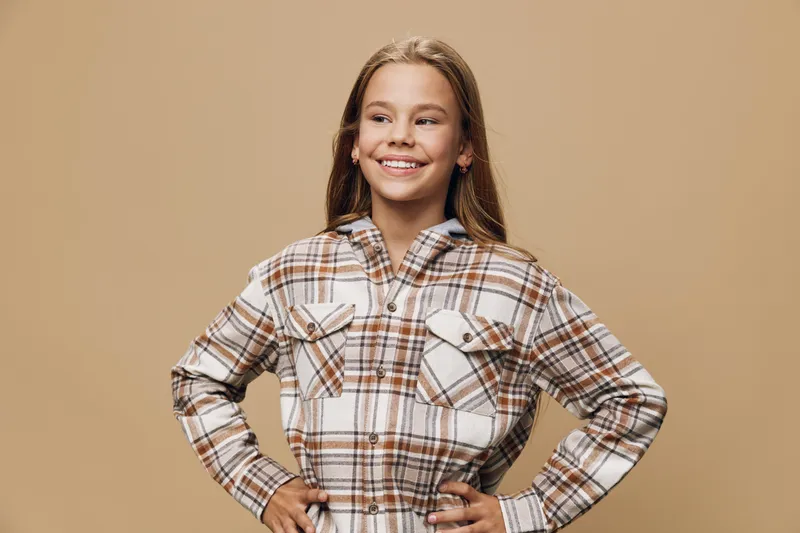 Smiling girl posing with hands on hips wearing plaid shirt against beige background looking confident and happy with casual style and youthful expression
