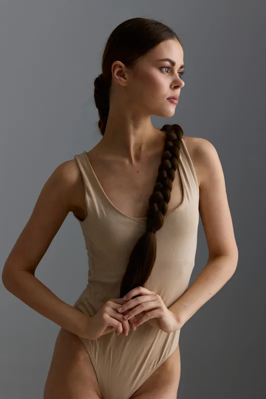 young woman with long braid in beige bodysuit stands against gray background, looking sideways with calm expression, minimalist fashion portrait in soft natural light