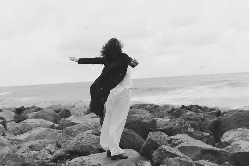 Woman stands on rocky coastline with arms outstretched, embracing wind and sea, monochrome scene of freedom, movement and exploration by the shore, calm mood outdoors