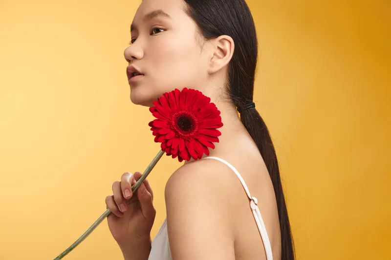 Asian woman holds a red flower near her face in a portrait against a warm yellow backdrop, conveying elegance, calm, and artistic serenity with subtle grace and timeless charm, and a gentle, poised