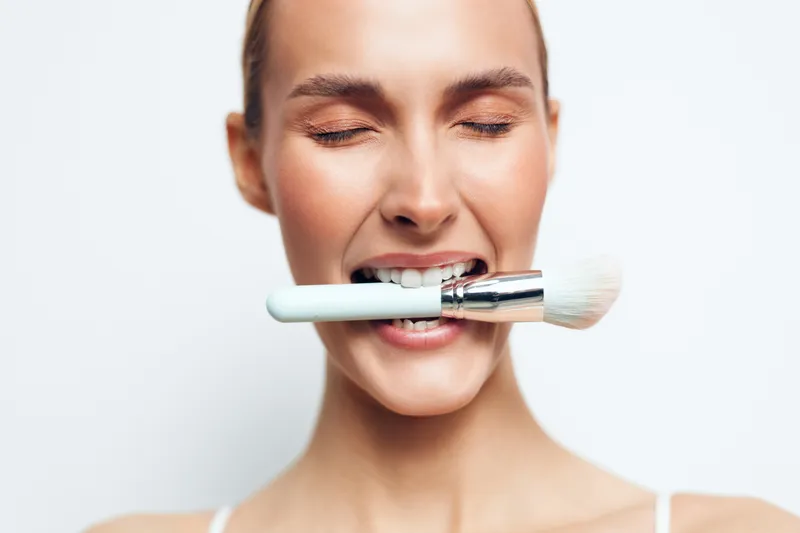 Playful young woman bites a makeup brush with eyes closed, bright studio light, clean background, casual pose, soft makeup, intimate beauty moment