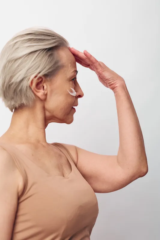 Side profile of mature woman applying cream on face with hand raised to forehead. Concept of skincare, aging, beauty routine, and self-care with soft natural lighting and neutral background.