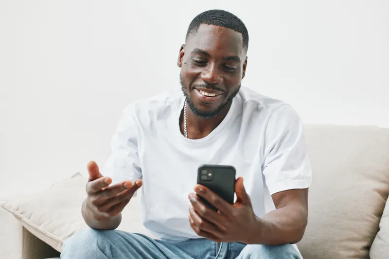 Smiling Black man in a casual white t shirt using a smartphone, showcasing engagement and happiness in a bright, minimalistic living room setting