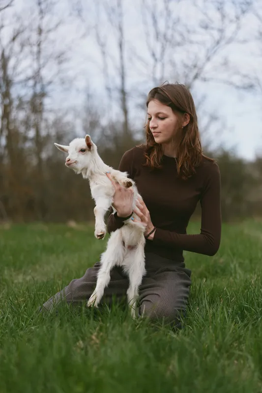 Young woman in a brown sweater gently holds a white baby goat while sitting on green grass in an outdoor natural setting during early spring.