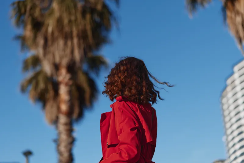 Woman with curly hair in a red jacket seen from behind, outdoors with palm trees and clear blue sky, urban fashion, sunny mood, fresh breeze and motion.