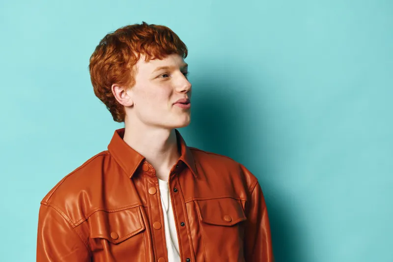 Young redhead man with curly hair wearing brown leather jacket and white shirt posing sideways on blue background looking thoughtful and confident in casual style portrait.