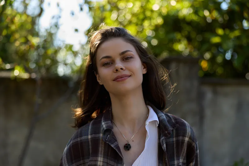 Young woman in a plaid shirt stands outdoors in a sunlit park, her calm smile and relaxed posture convey casual confidence and natural lifestyle