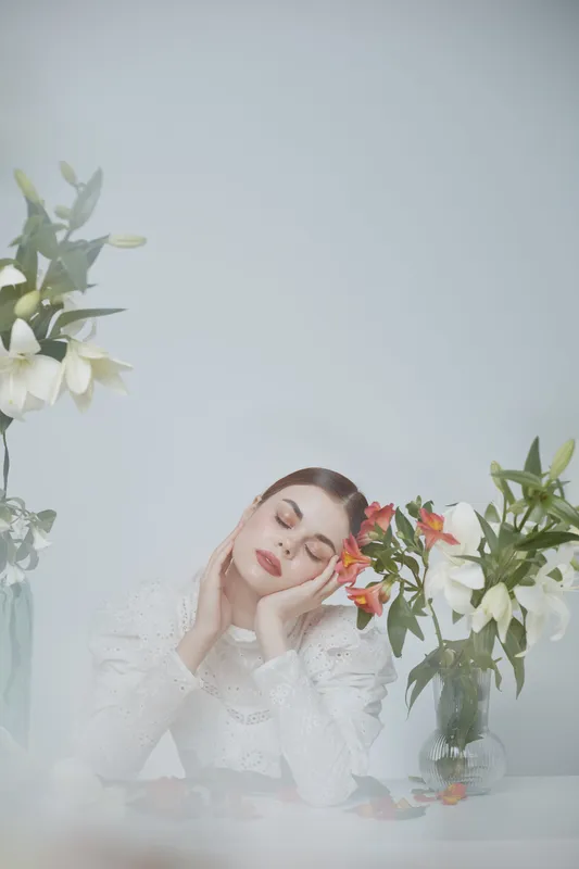 A serene young woman in white attire surrounded by fresh flowers, exuding calmness and tranquility in a soft, floral setting