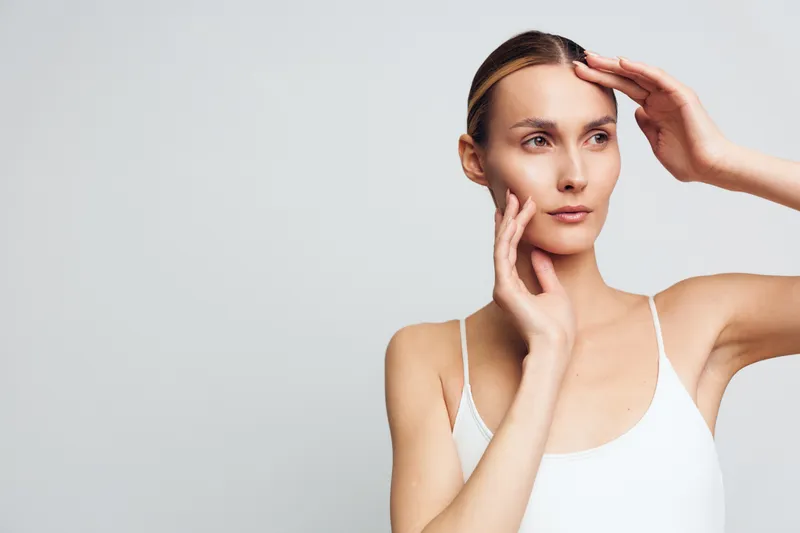 Focused woman in a white tank top touches her forehead with one hand and rests her chin on the other, isolated on a neutral background, conveying concentration and subtle elegance.
