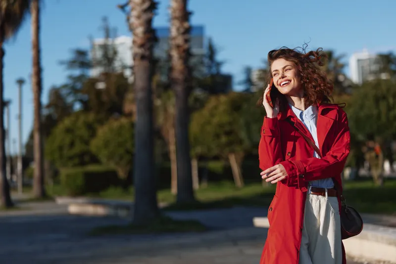 Young woman in a red coat stands in a sunny park, talking on a mobile phone, smiling as a light breeze moves her hair in a candid outdoor moment